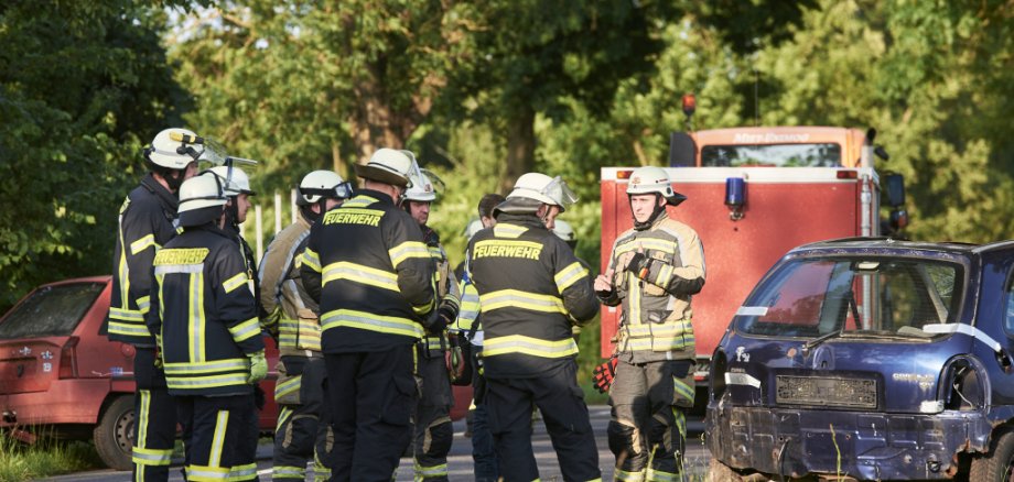 The picture shows several firefighters in turnout gear during an exercise in front of a car involved in an accident. Apart from one firefighter, all the other people can only be seen from behind. There is a car on the left and right-hand side of the road and a fire engine in the background.